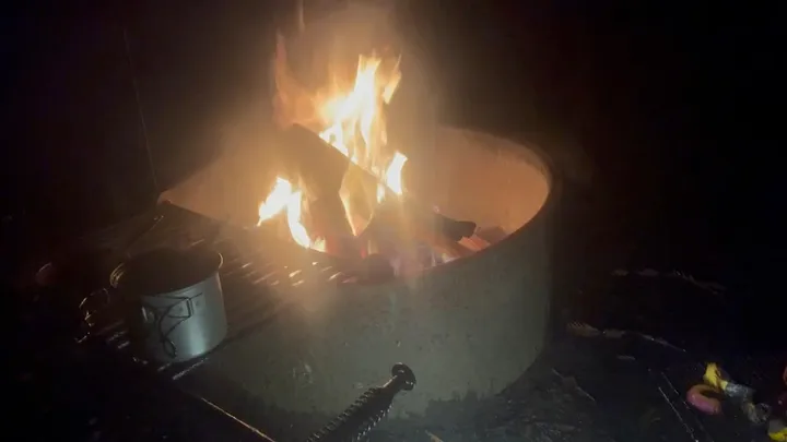 A campfire burning in a fire ring at night, with a camp mug on the grill.